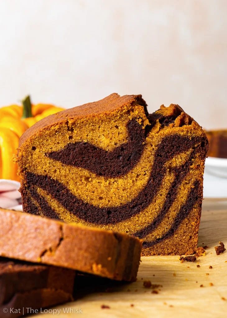 Gâteau marbré à la citrouille sans gluten, avec quelques tranches déjà coupées, sur une feuille de papier sulfurisé marron, avec deux petites citrouilles en arrière-plan.