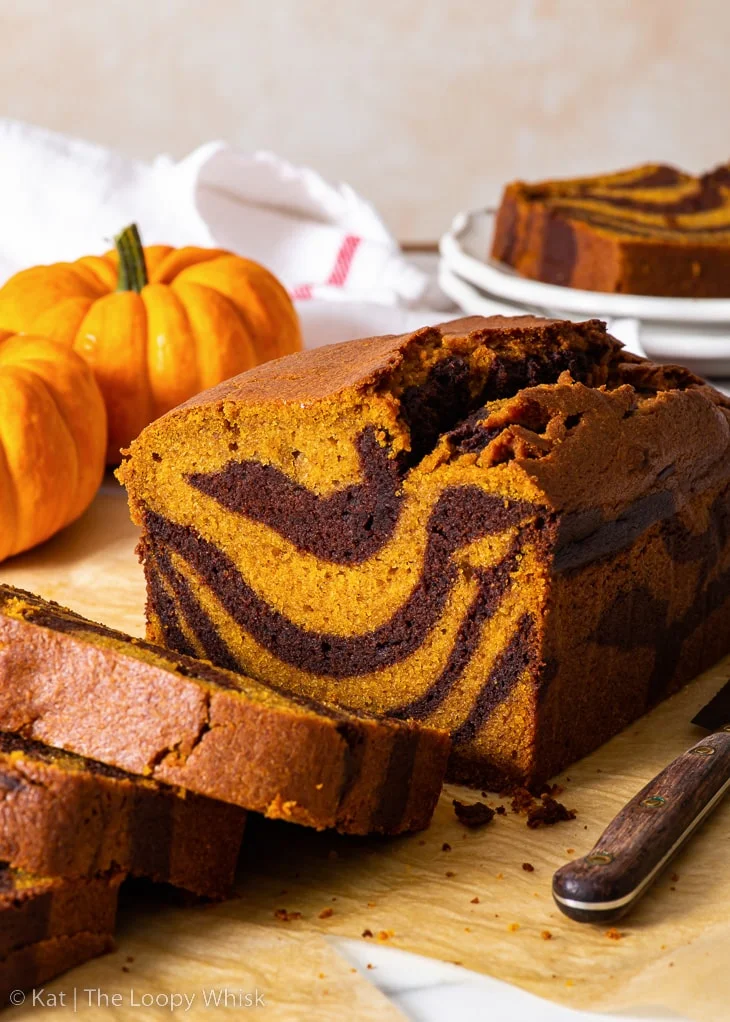 Gâteau marbré à la citrouille sans gluten, avec quelques tranches déjà coupées, sur une feuille de papier sulfurisé marron, avec deux petites citrouilles en arrière-plan.